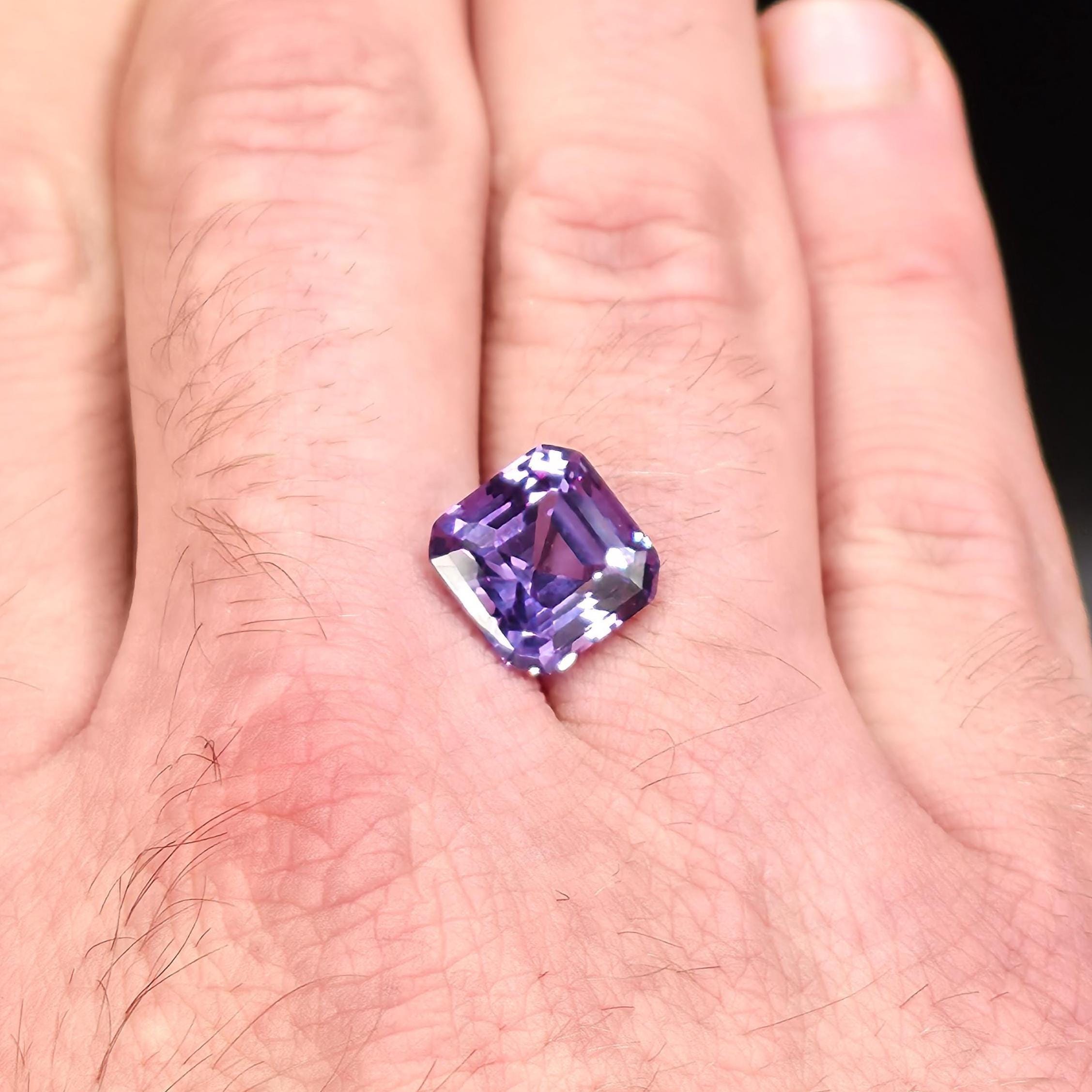 Emerald-cut Aetherium Alexandrite resting on hand, displaying intense color shift from violet-blue to pinkish tones. High-clarity synthetic alexandrite gemstone for fine jewelry or collector display, precision-cut from Aetherium rough material.