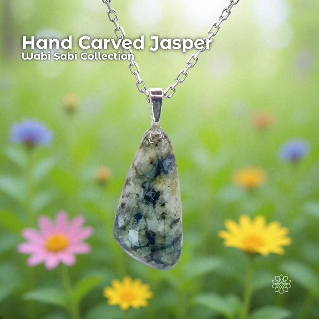 A hand-carved jasper pendant set in a sterling silver bail, hanging from a delicate silver chain against a soft-focus garden background. The stone is an organic triangular shape with natural cream, green, and dark inclusions. Text on the image reads “Hand Carved Jasper – Wabi Sabi Collection,” with a small Aetherium Daisy Logo in the lower right corner.