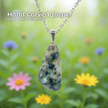 A hand-carved jasper pendant set in a sterling silver bail, hanging from a delicate silver chain against a soft-focus garden background. The stone is an organic triangular shape with natural cream, green, and dark inclusions. Text on the image reads “Hand Carved Jasper – Wabi Sabi Collection,” with a small Aetherium Daisy Logo in the lower right corner.