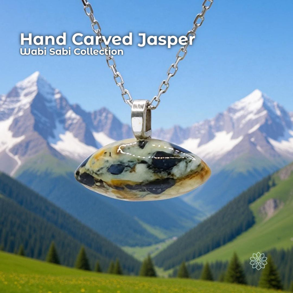 A hand-carved jasper cabochon pendant set in a sterling silver bail, hanging from a delicate silver chain with snow-covered mountains and a bright green valley in the background. The stone is oval with natural cream, black, and golden markings. Text on the image reads “Hand Carved Jasper – Wabi Sabi Collection,” with a small Aetherium floral mark in the lower right corner.