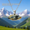 A hand-carved jasper cabochon pendant set in a sterling silver bail, hanging from a delicate silver chain with snow-covered mountains and a bright green valley in the background. The stone is oval with natural cream, black, and golden markings. Text on the image reads “Hand Carved Jasper – Wabi Sabi Collection,” with a small Aetherium floral mark in the lower right corner.