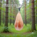 A hand-carved rose quartz pendant set in a sterling silver bail, hanging from a delicate silver chain in a soft green forest background. The stone is an organic teardrop shape with pale pink and warm reddish tones. Text on the image reads “Hand Carved Quartz – Wabi Sabi Collection,” with a small Aetherium floral mark in the lower right corner.