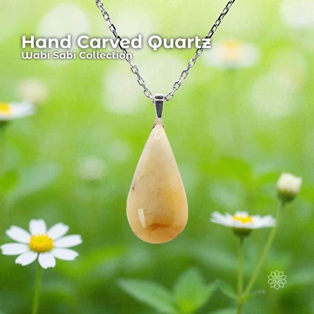 A hand-carved golden quartz pendant set in a sterling silver bail, hanging from a delicate silver chain against a soft green garden background with small white and yellow flowers. The stone is an elongated teardrop shape with warm honey and amber tones. Text on the image reads “Hand Carved Quartz – Wabi Sabi Collection,” with a small Aetherium floral mark in the lower right corner.