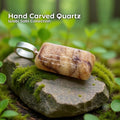 A hand-carved orange aventurine quartz pendant set in a sterling silver bail, resting on a moss-covered stone in a forest setting. The quartz is a polished rectangular shape with warm orange, cream, and subtle marbled patterns. Text on the image reads “Hand Carved Quartz – Wabi Sabi Collection,” with a small Aetherium floral mark in the lower right corner.