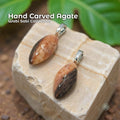 A pair of hand-carved two-tone agate pendants set in sterling silver bails, photographed against a forest background. Each stone is an elongated oval with warm , cream, and dark brown banding. Text on the image reads “Hand Carved Agate – Wabi Sabi Collection,” with a small Aetherium floral mark in the lower right corner.