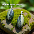 Two hand-carved green banded jasper pendants set in sterling silver bails, hanging from silver chains against a blurred forest background. The stones are elongated oval shapes with natural dark and light green banding. Text reads “Hand Carved Jasper – Wabi Sabi Collection,” with a small Aetherium floral mark in the lower right corner.