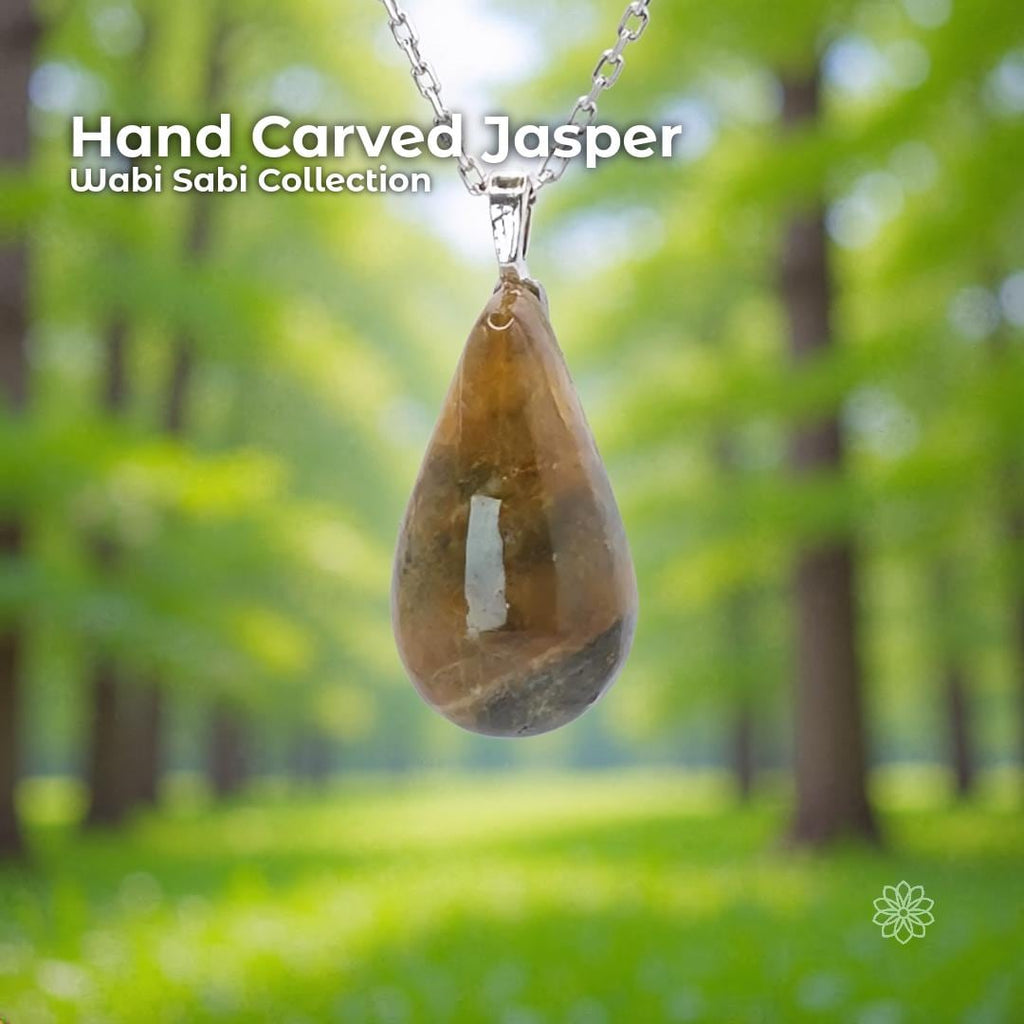 A hand-carved banded jasper pendant set in a sterling silver bail, hanging from a silver chain against a soft green forest background. The stone is a polished teardrop shape with natural warm brown, amber, and gray banding. Text on the image reads “Hand Carved Jasper – Wabi Sabi Collection,” with a small Aetherium floral mark in the lower right corner.