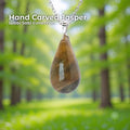 A hand-carved banded jasper pendant set in a sterling silver bail, hanging from a silver chain against a soft green forest background. The stone is a polished teardrop shape with natural warm brown, amber, and gray banding. Text on the image reads “Hand Carved Jasper – Wabi Sabi Collection,” with a small Aetherium floral mark in the lower right corner.