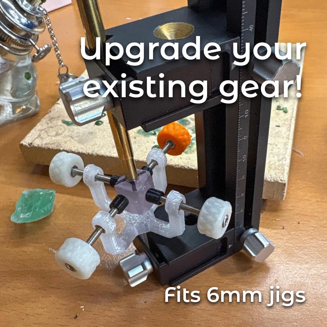 Gem Axis V3 attached to a black faceting mast with a brass quill; the clear recycled-polycarbonate frame, white knurled wheels, and an orange knob center a 6 mm dop. Rough green stone pieces and bench debris sit nearby, indicating active cutting. On-image text: “Upgrade your existing gear! • fits 6mm jigs.” Shows professional-grade, beginner-friendly realignment to restart cuts accurately.
