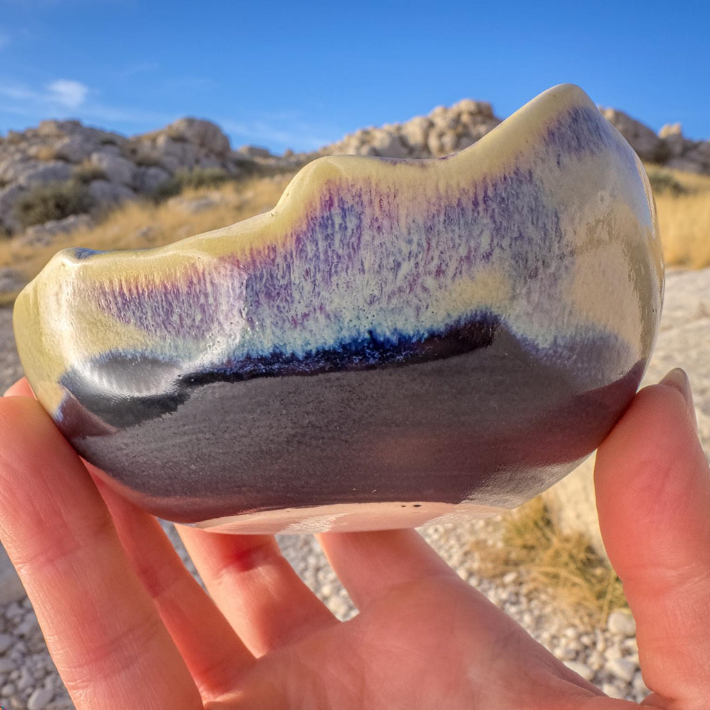 Side view of the Erosion Study No. 1 handmade ceramic dish held outdoors. Glaze transitions from creamy yellow to deep indigo and black with violet undertones, emphasizing sculpted edges inspired by eroded stone. Captured in natural desert light for the Wabi Sabi Collection by Aetherium Innovations.