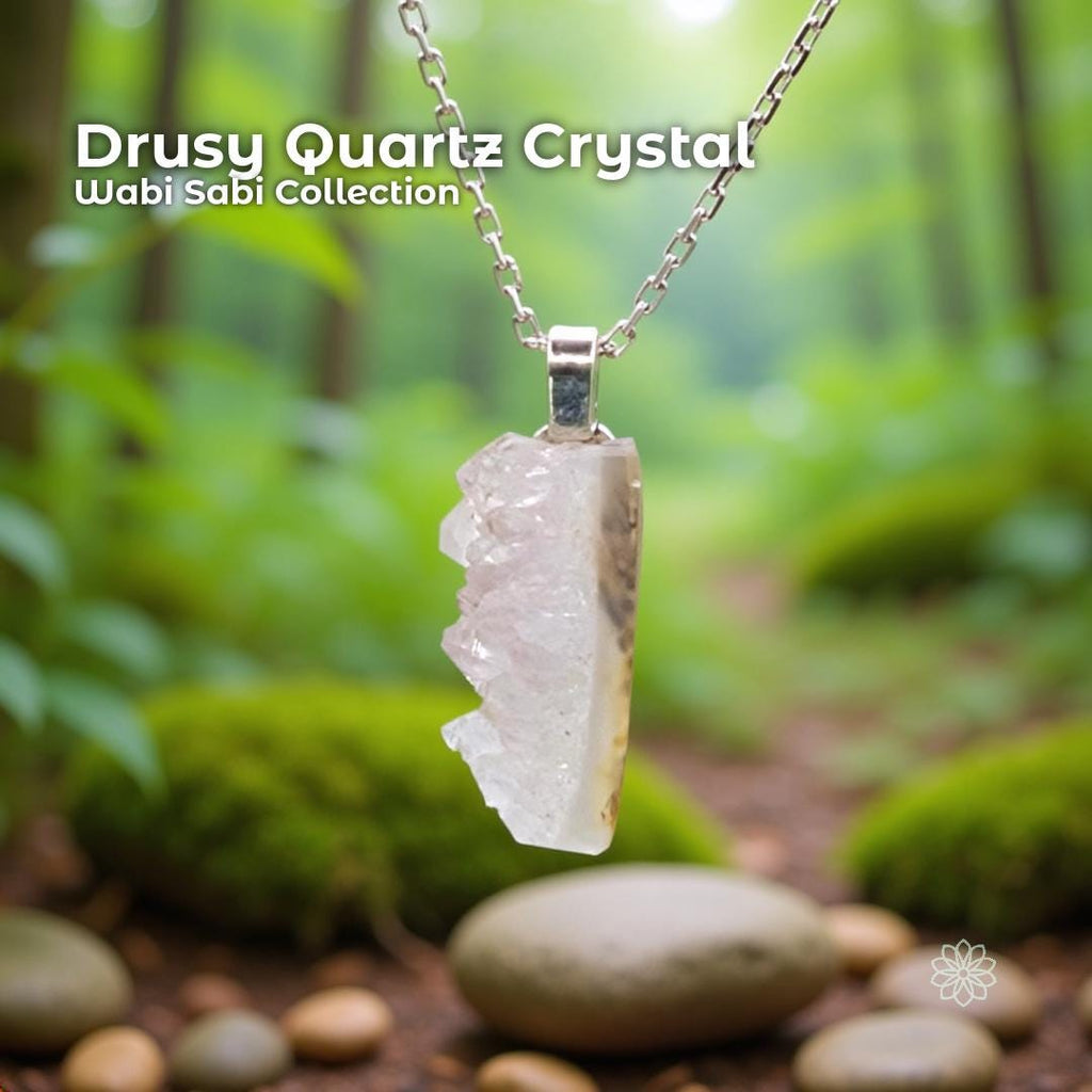 A raw drusy quartz crystal pendant with sparkling natural crystal texture set in a sterling silver bail. The stone hangs in front of a soft green forest scene with moss, river rocks, and blurred trees. Part of the Wabi Sabi Collection, it highlights organic shape, earthy tones, and Pacific Northwest handcrafted style.