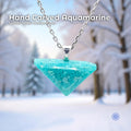 Hand carved aquamarine pendant in a triangular polished shape, hanging from a sterling silver chain. The stone is bright icy blue with natural marbling. The background shows a soft snowy forest with trees and winter light, highlighting the nature-inspired, handmade Pacific Northwest style.