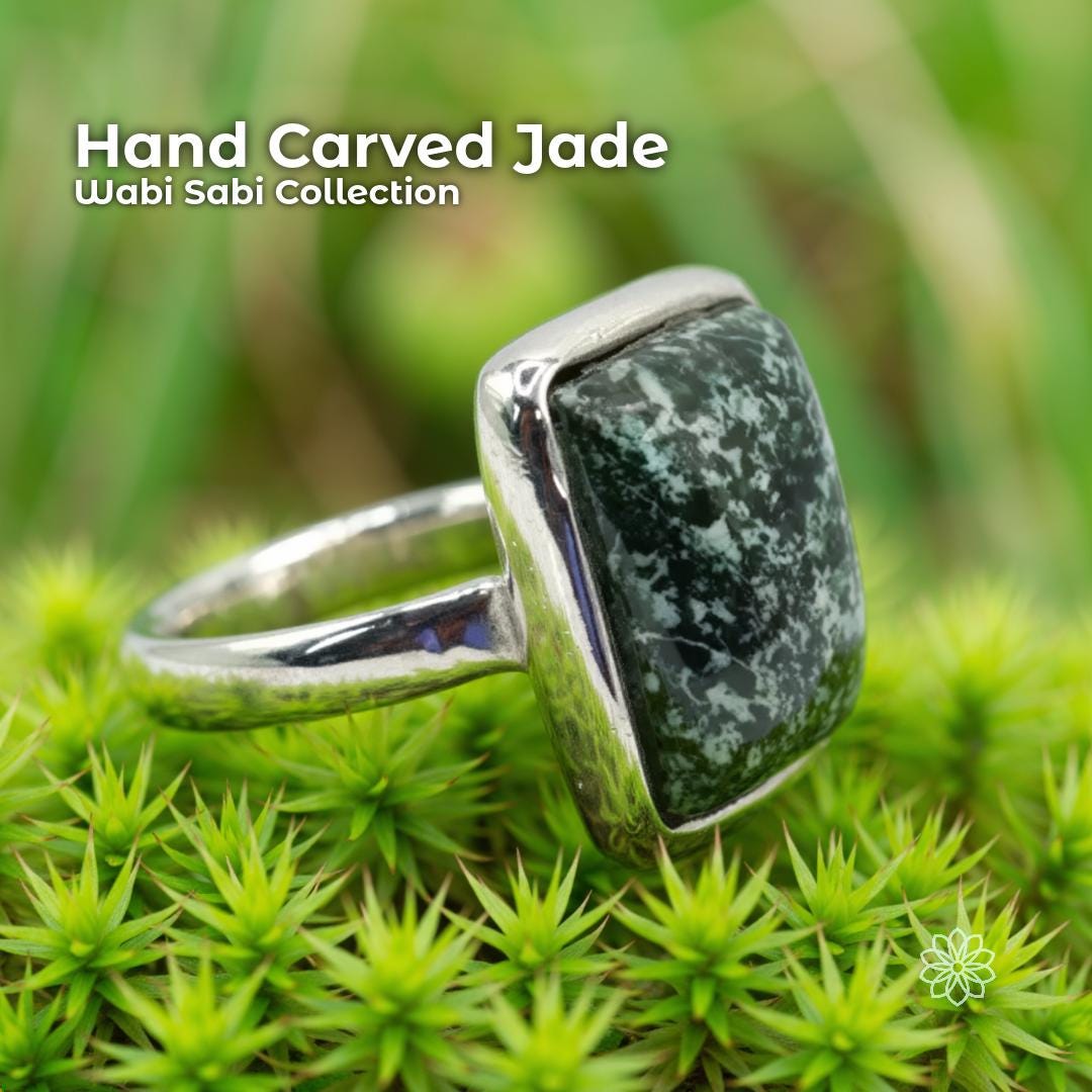 Hand carved nephrite jade ring with a dark green stone featuring white speckled patterns, set in a solid sterling silver band. The ring is photographed resting on bright green moss with soft blurred background foliage, emphasizing its natural, earthy, Pacific Northwest style.
