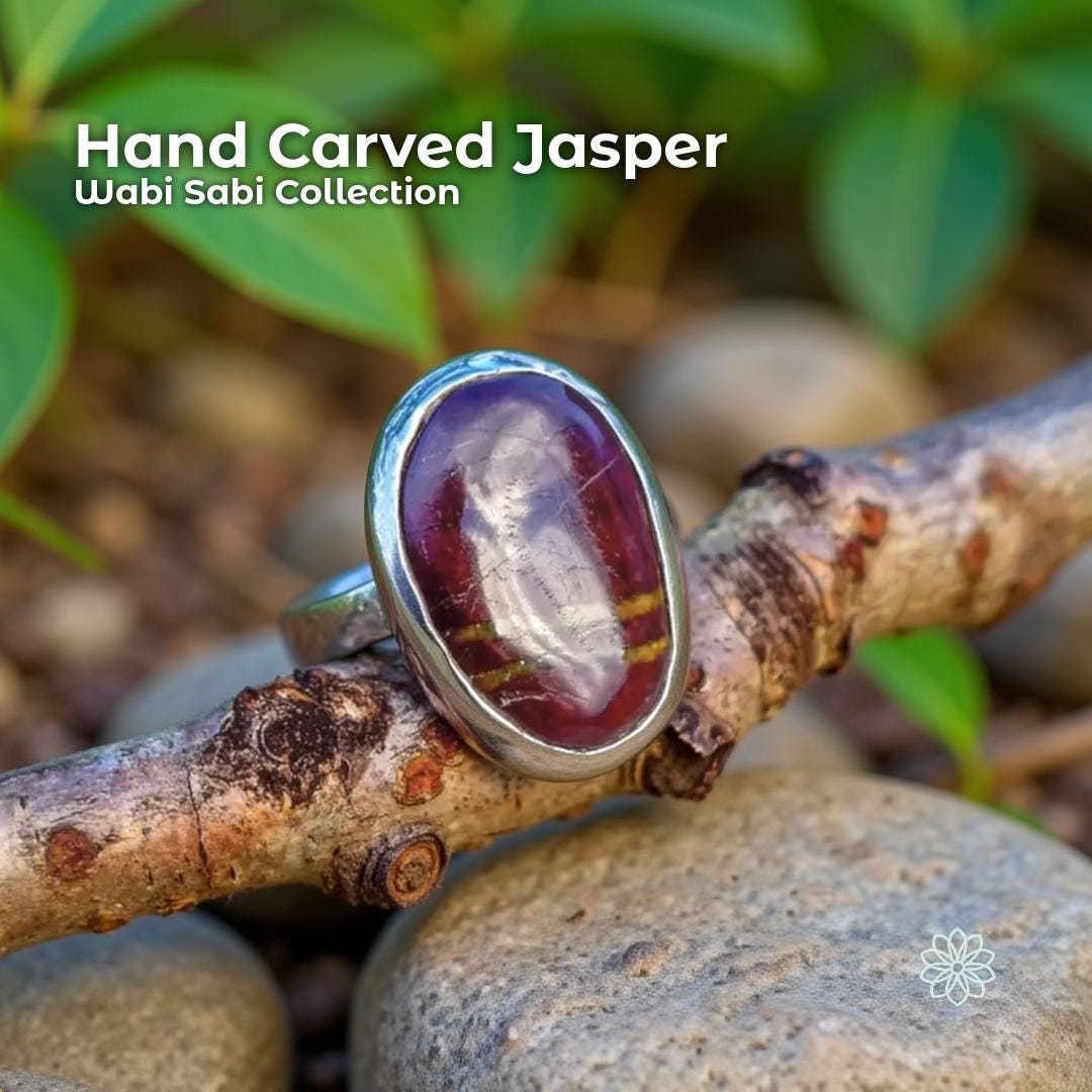 Hand carved red banded jasper ring set in a sterling silver band. The oval jasper stone shows deep red tones with natural striping and a raw, unsealed finish. The ring is photographed resting on a twig among smooth river stones and green leaves, creating an earthy, nature-focused Pacific Northwest aesthetic.