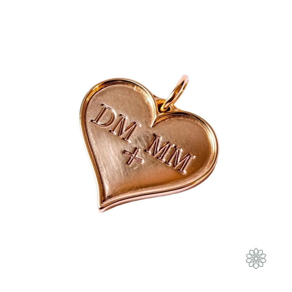 Heart-shaped pendant engraved with the initials “DM + MM,” cast in warm gold with a polished surface and soft beveled edge. The charm rests on a white background, capturing fine metallic reflection and subtle engraving detail.
