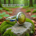 A handcrafted yellow quartz cabochon ring set in polished sterling silver resting on moss-covered stone in a forest setting, showing its warm golden translucence, natural inclusions, rounded dome cut, and textured band.