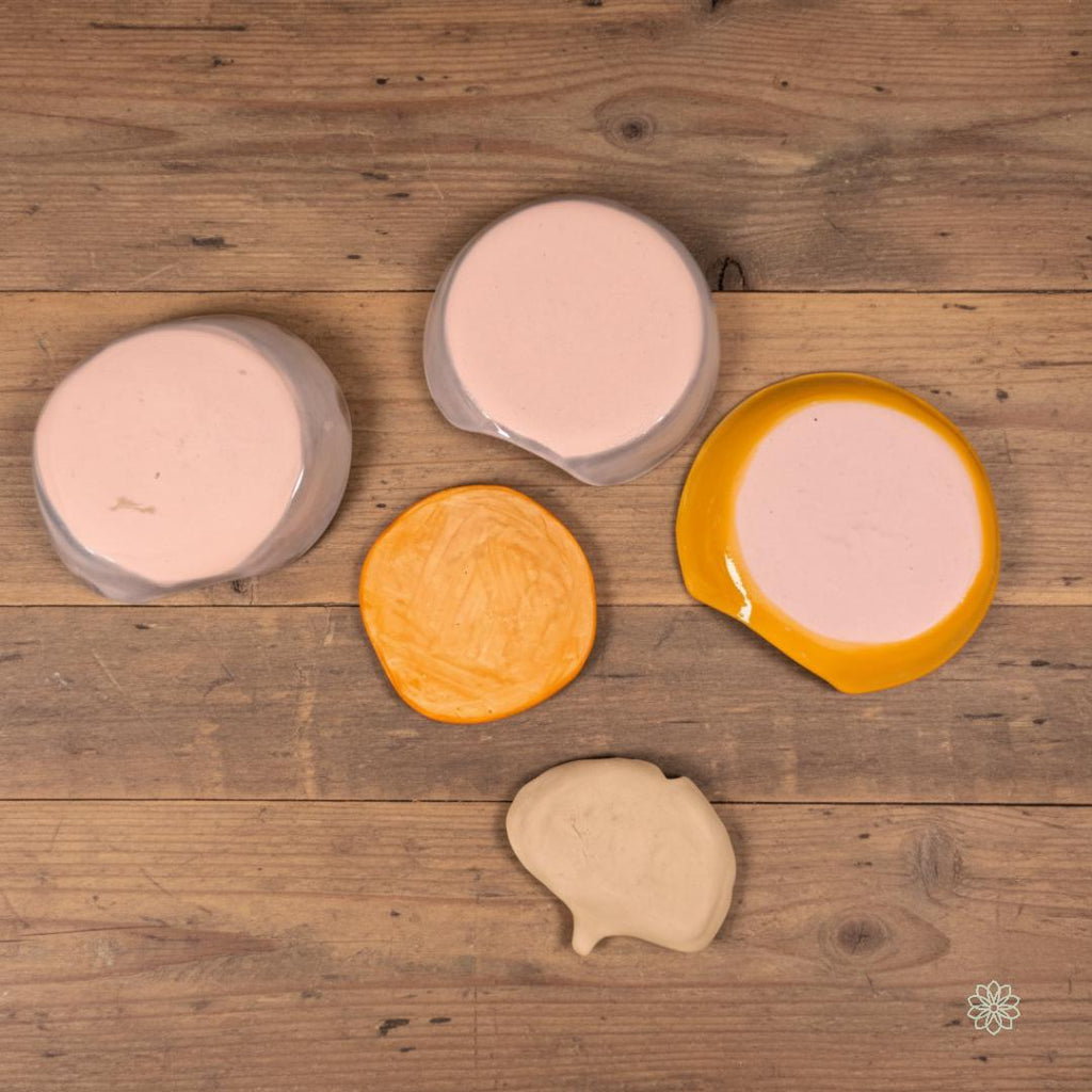 The underside view of five handmade ceramic dishes arranged on a rustic wooden surface, showing unglazed bases, clay body textures, and natural maker variations. Shapes include circular, organic, and leaf forms. Highlights craftsmanship, kiln-fired finish, and authenticity of the Aetherium × Archibald artisan pottery line.