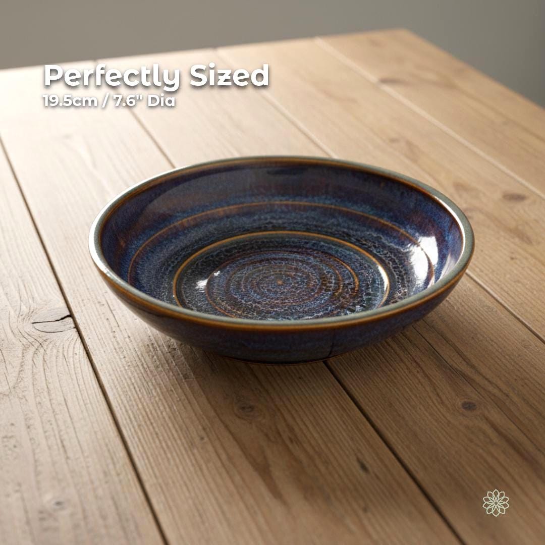 “Angled view of a handmade stoneware pasta bowl measuring 19.5 cm (7.6 inches) in diameter. The reactive glaze blends blue, rust, and gold tones, with a smooth glossy interior and thick ceramic walls. Shown on a light farmhouse wood table for size reference.”