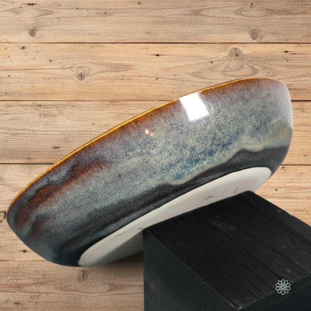 “Close-up side detail of the handmade stoneware pasta bowl, featuring a reactive glaze with layered amber, denim blue, and charcoal tones. The curved wall and glossy finish highlight the bowl’s craftsmanship. Photographed on a wood surface from the Aetherium × Archibald line.”
