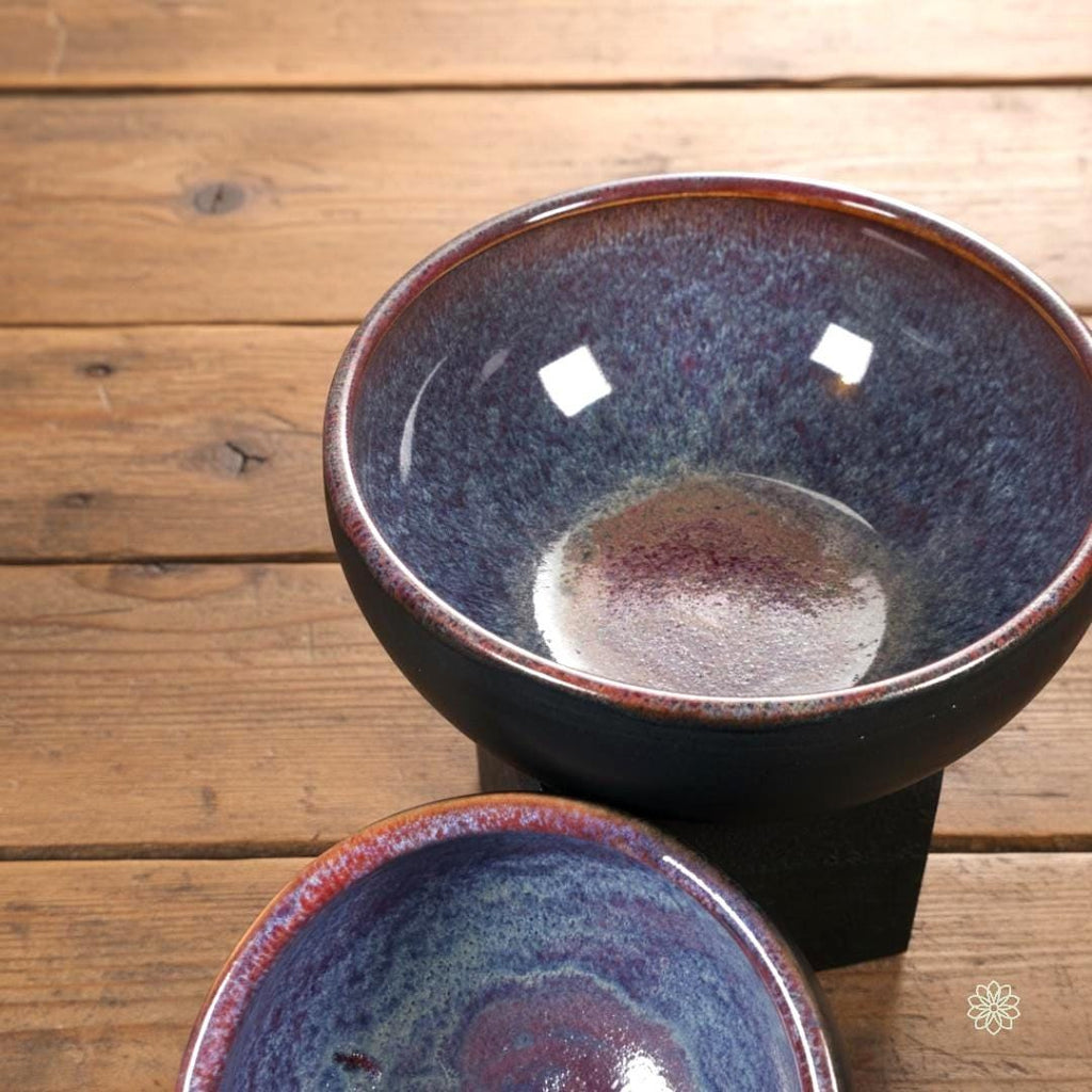 “Close-up of a large handmade stoneware bowl featuring a reactive glaze interior with a magenta center fading into blue and violet crystalline patterns. Glossy surface with light reflections. Shown on a warm wood table.”