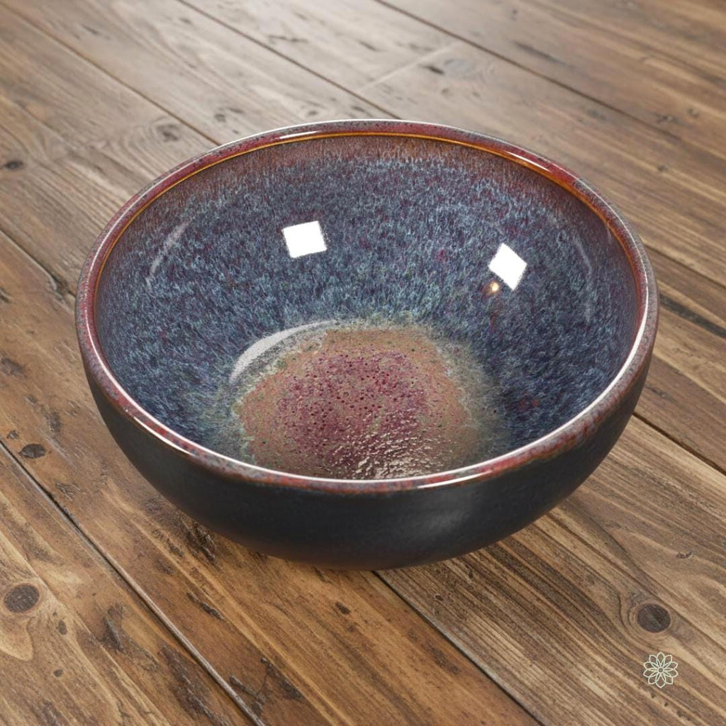 “Handcrafted stoneware bowl with a reactive glaze interior blending magenta, violet, and blue tones. The bowl is shown at an angle to emphasize depth and curved form. Glossy interior, matte black exterior, on a rustic wood surface.”