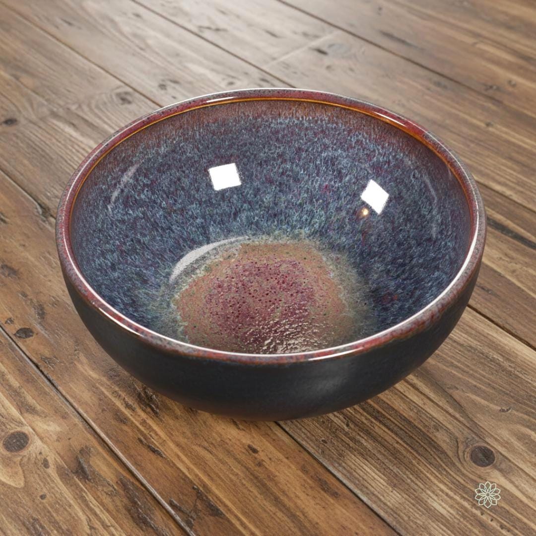 “Handcrafted stoneware bowl with a reactive glaze interior blending magenta, violet, and blue tones. The bowl is shown at an angle to emphasize depth and curved form. Glossy interior, matte black exterior, on a rustic wood surface.”
