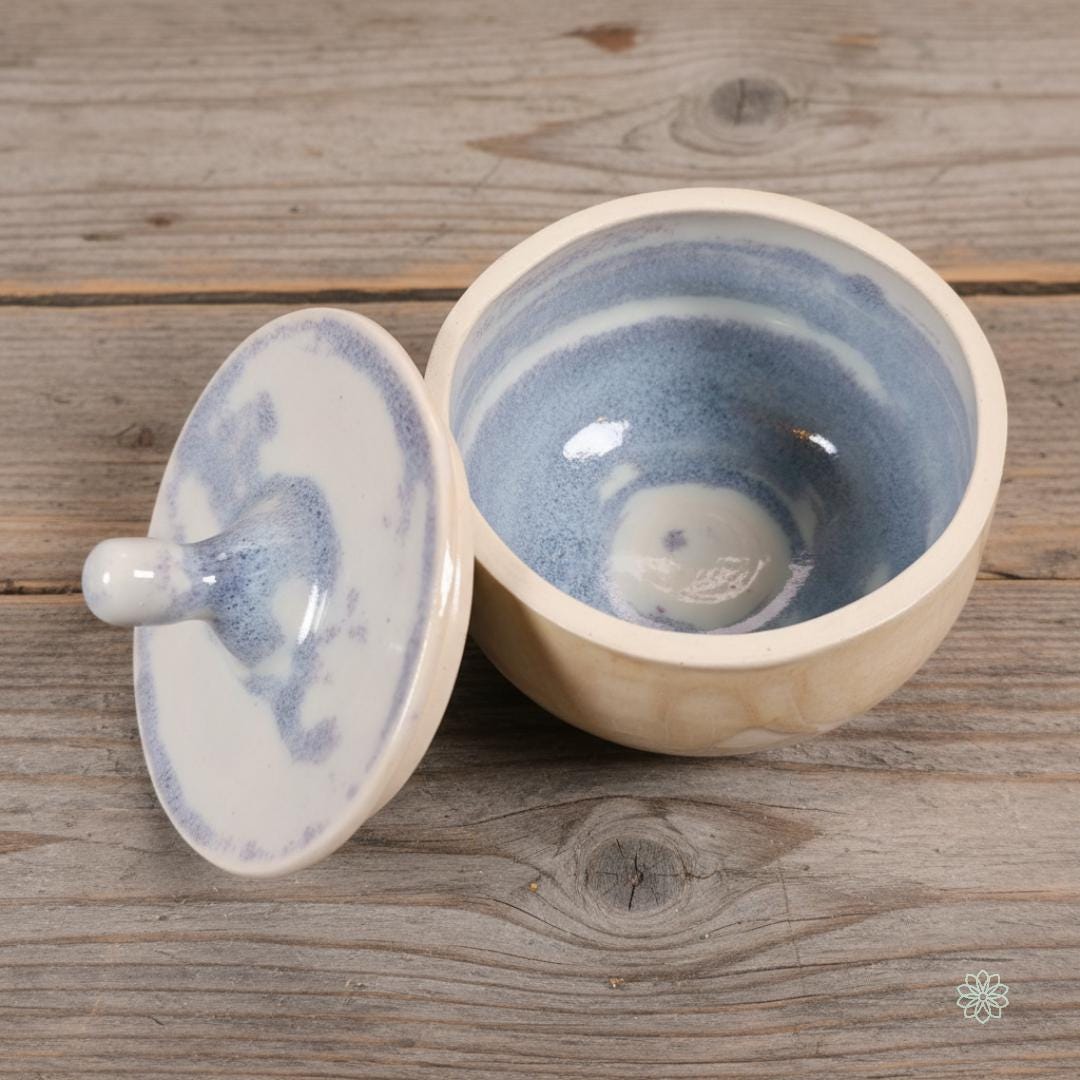 “White and blue marbled ceramic altar jar with the lid placed beside the open bowl, showing glossy swirl glazing inside. Handmade pottery styled on rustic grey wood. Part of the Aetherium x Archibald altar jar set.”