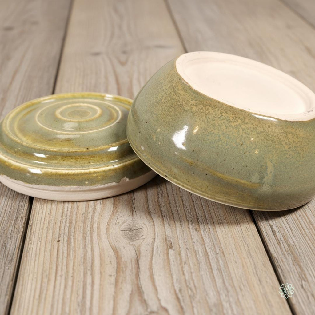 “Open green ceramic altar jar displayed on light farmhouse wood, showing the lid with carved concentric rings beside the matching bowl base. Handmade pottery with soft green glaze and cream stoneware foot. Part of the Aetherium x Archibald ceramic altar collection.”