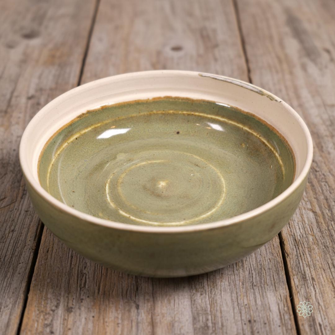 A small handmade ceramic altar jar in soft olive green glaze shown with the lid removed. The lid displays raised concentric rings; the base shows a smooth interior and pale unglazed foot. Both pieces rest on light farmhouse-style wood. This jar is labeled “Green Jar” in other product photos and measures approximately 8 cm / 3 inches in diameter.