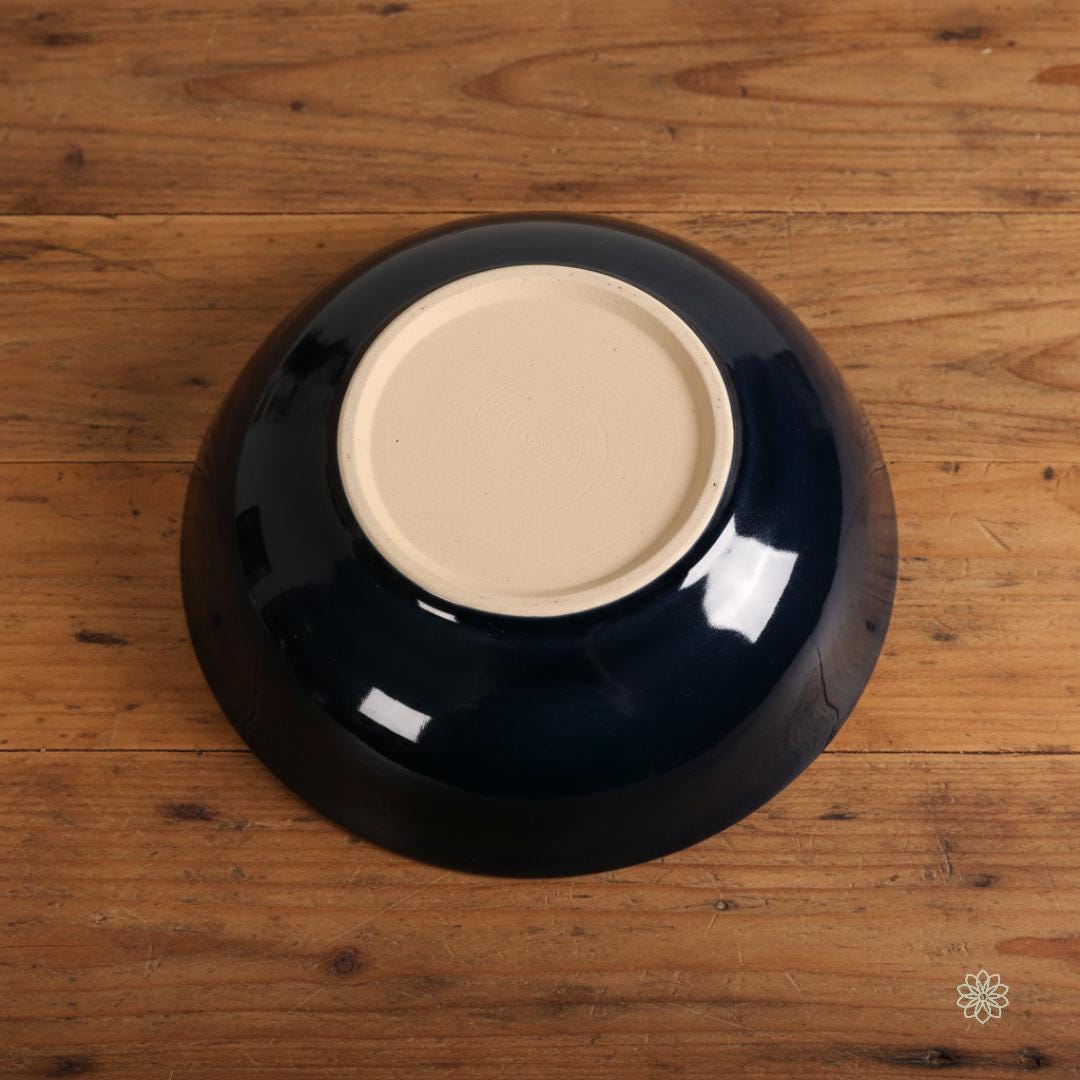 Bottom view of a reactive glaze stoneware bowl resting on a wooden surface. The underside is fully glazed in dark navy blue with a glossy sheen, while the circular foot ring is unglazed light stoneware clay, showing trimmed throwing lines. No printed text or decoration appears on the base.