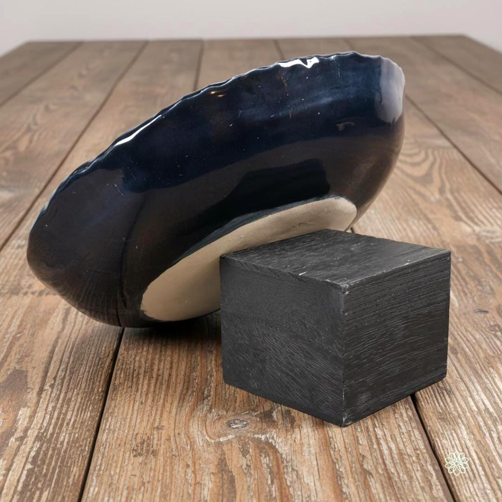 Side view of a large braided-rim stoneware bowl propped on a dark wooden block to reveal its profile. The outside of the bowl is glazed in glossy dark blue. The unglazed tan stoneware foot ring is partly visible, showing how the bowl sits on a table. Wood plank surface in the background.
