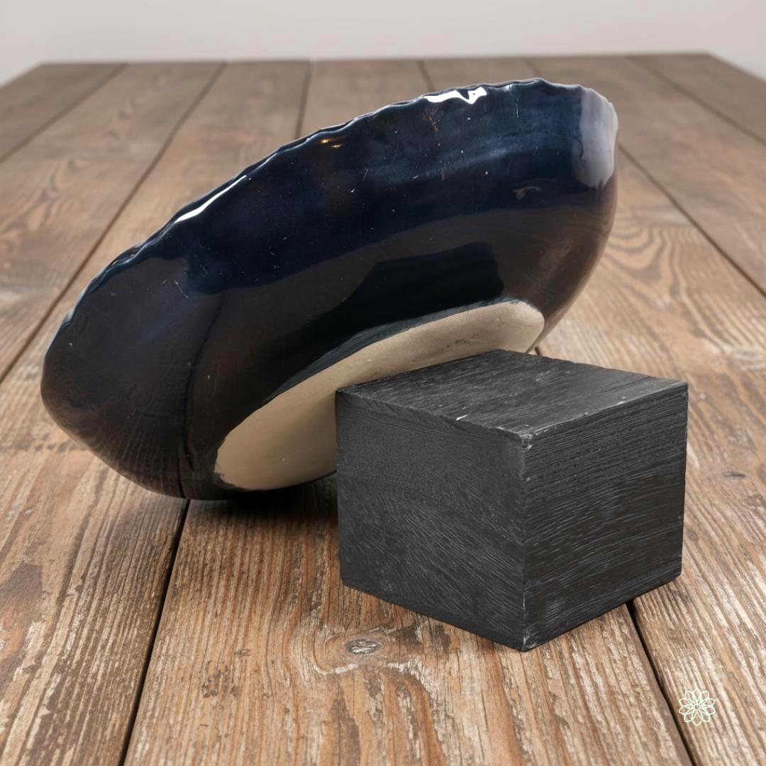 Side view of a large braided-rim stoneware bowl propped on a dark wooden block to reveal its profile. The outside of the bowl is glazed in glossy dark blue. The unglazed tan stoneware foot ring is partly visible, showing how the bowl sits on a table. Wood plank surface in the background.