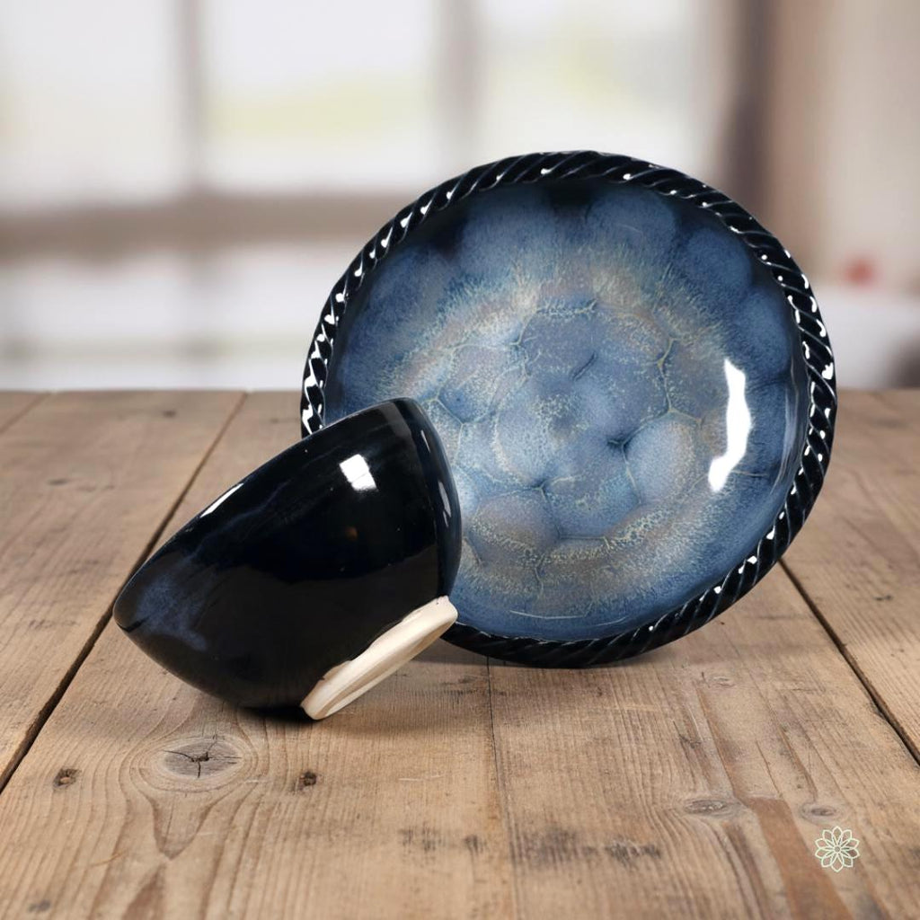 Two reactive glaze stoneware bowls displayed on a wooden table. The large braided-rim bowl stands upright to show a blue crystalline interior pattern, while the smaller bowl rests at an angle in front, showing its dark blue exterior and unglazed foot. Background is softly blurred to suggest an indoor home setting.