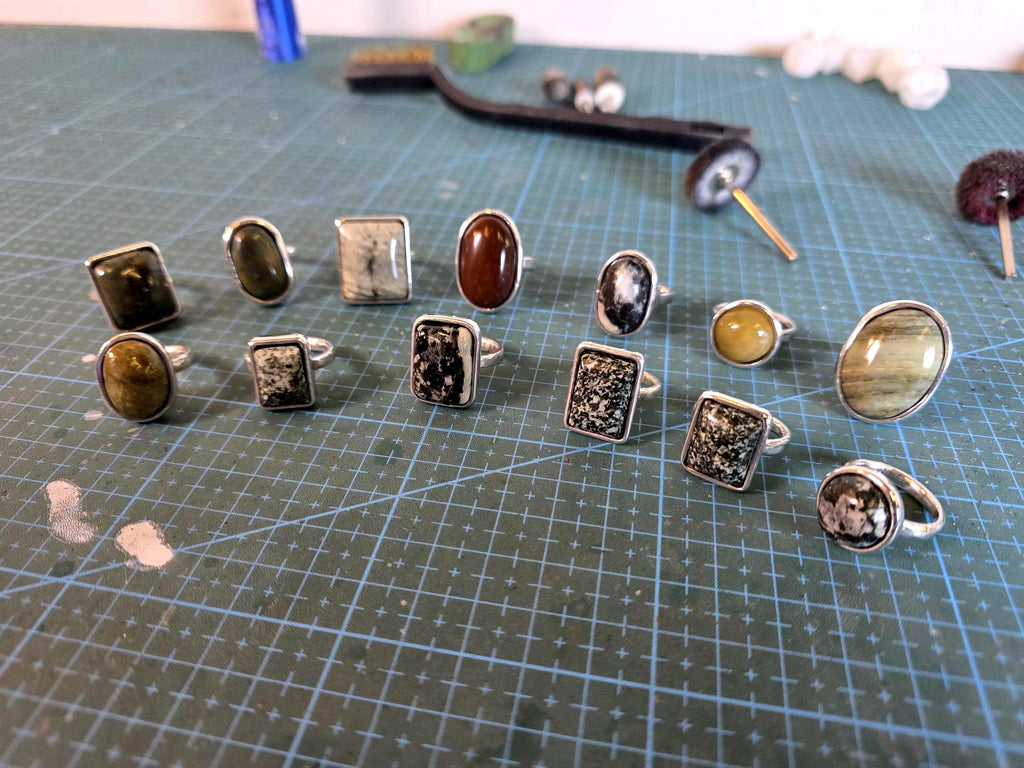 Photo of finished cabochon rings in silver, featuring oval and rectangular settings with natural stones. Displayed on a cutting mat with polishing tools in the background, showing final polish and craftsmanship achievable from the printed blanks.