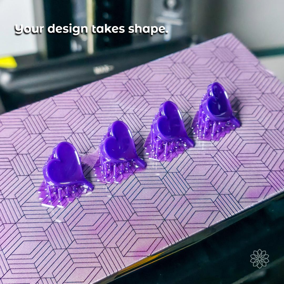Row of purple resin heart models freshly printed on a textured build plate, showing the 3D printing phase of custom jewelry production. Demonstrates prototype creation, precision modeling, and readiness for lost-wax casting. Ideal for clients curious about how their concept becomes a physical form.