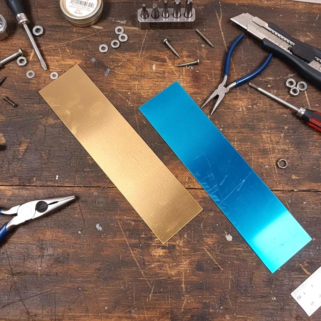 Two 2x8 inch brass engraving blanks shown side by side on a wood workbench. The left sheet is uncovered, revealing its golden raw brass finish with a slight reflective sheen. The right sheet still has its protective blue film intact, displaying surface texture and light scuffing. Both sheets are 18 gauge, 1mm thick, with square corners and flat-cut edges. Ideal for CNC engraving, custom plaques, jewelry tags, nameplates, and personalized logo work.