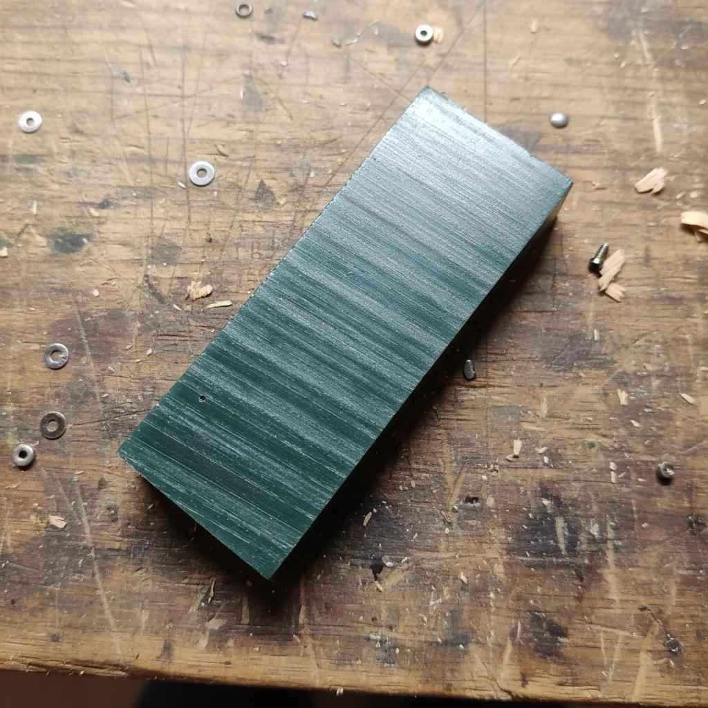 “A rectangular block of green carving wax resting on a workbench. The wax shows uniform machining marks along the surface, indicating a clean mill pass suitable for CNC modeling, hand-carving, jewelry patterns, or lost-wax casting prototypes. Small metal shavings and tools surround the block.”