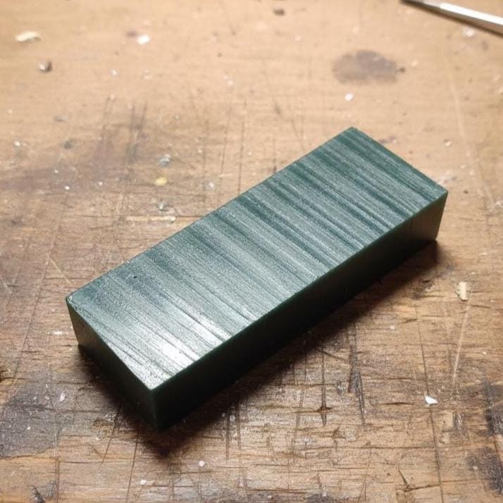 “A machined green carving-wax blank on a jeweler’s bench, with textured tool-path lines visible across the surface. This wax slab is used for ring carving, CNC machining, sculpting small jewelry forms, and preparation for investment casting. Loose hardware and debris sit around the workspace.”