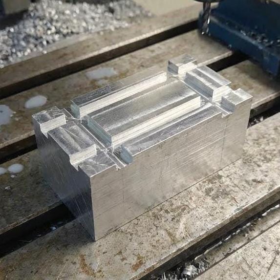 Machined 50×50×50 mm 6061-T6 aluminum block shaped into a stepped form on a CNC bed with chips around it. Smooth toolpaths and crisp features. Suitable for mold making, machining practice, test cuts, precision fixturing, and CNC learning material.