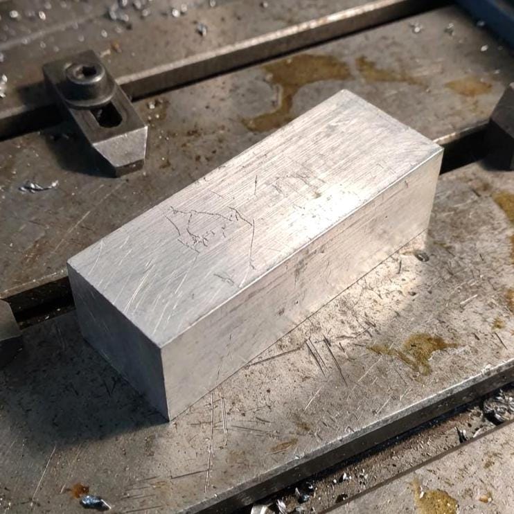 50×50×50 mm 6061-T6 aluminum block sitting on a milled steel bed with coolant stains and scattered aluminum chips. Even machined surface with sharp edges. Ideal CNC bar stock for milling setups, workholding tests, custom parts, prototyping, and metal shop projects.