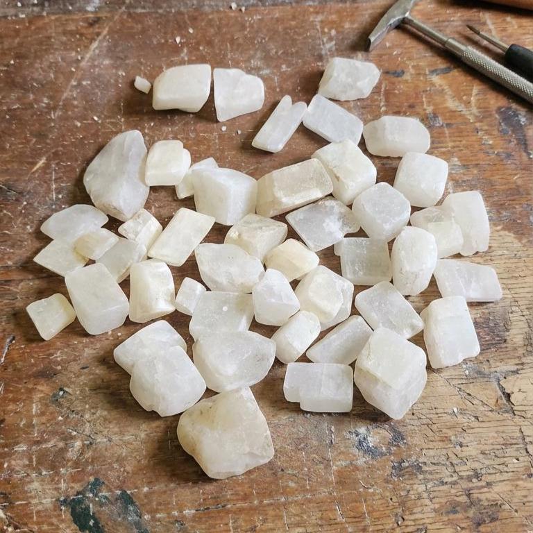 A large pile of lightly tumbled calcite pieces displayed outdoors on weathered granite and quartz-bearing matrix. The stones range from soft pink to pale yellow with semi-translucent structure. Natural daylight highlights internal cleavage planes and surface texture, making the material suitable for cabbing, carving, inlay work, and geology instruction.