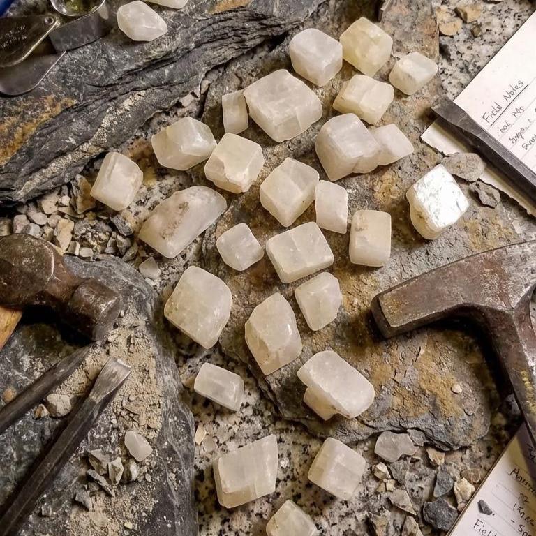 A group of pale calcite rough chips arranged among rock slabs, field tools, and geological notebooks. Hammer marks and scattered fragments show active specimen preparation. The calcite pieces have square and rectangular cleavage faces and are sized for lapidary study, inlay work, or geology teaching. Scene presents the material in a realistic field-lab hybrid setting.