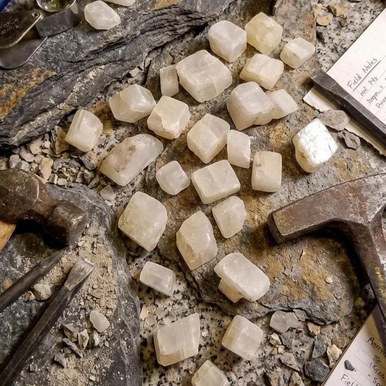A group of pale calcite rough chips arranged among rock slabs, field tools, and geological notebooks. Hammer marks and scattered fragments show active specimen preparation. The calcite pieces have square and rectangular cleavage faces and are sized for lapidary study, inlay work, or geology teaching. Scene presents the material in a realistic field-lab hybrid setting.