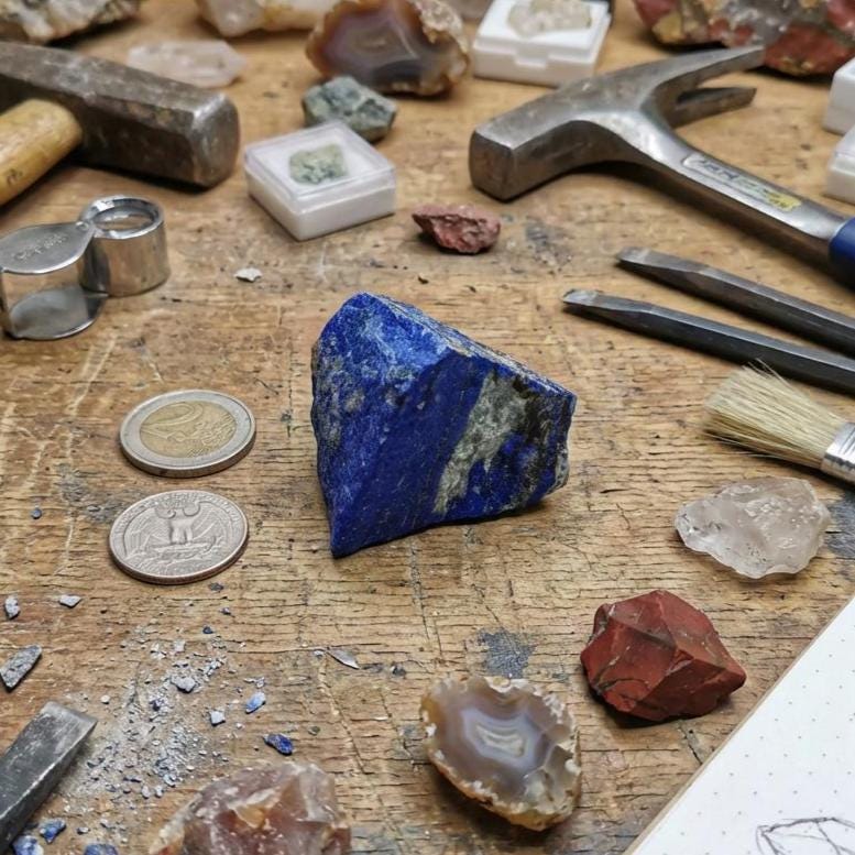 “A rough triangular piece of lapis lazuli showing bright blue lazurite, calcite veining, and pyrite speckling, displayed on a workbench with rock tools, agate slices, quartz points, and magnification tools. Scene evokes a lapidary studio environment for cutting, shaping, carving, or pigment grinding.”