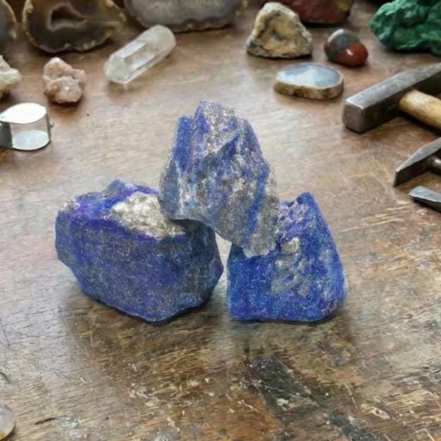 “Three rough lapis lazuli chunks stacked on a workbench, showing saturated ultramarine blue with patches of calcite and pyrite. Surrounding them are agates, quartz, chisels, a loupe, and lapidary debris. The composition reflects active mineral analysis and preparation for cutting, carving, or pigment extraction.”