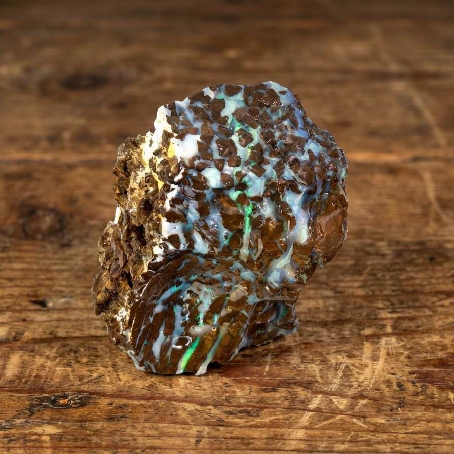 A large piece of Australian Boulder Opal rough on a wooden bench, showing blue, green, and white opal seams running through dark brown ironstone. The surface has natural cavities and ridges with visible fire. Ideal for lapidary cutting, cabbing, and seam-tracking practice. Raw, untreated material from Queensland mining fields.