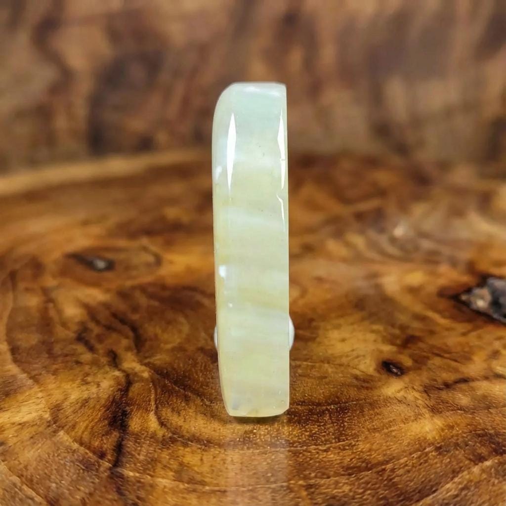 “A polished teardrop-shaped chalcedony agate lying on a textured wooden surface. The stone shows soft translucent whites and warm yellow tones with fine volcanic sand inclusions sealed inside the material. Gentle internal banding becomes visible where light reflects across the surface. A ghostly PNW agate formed from ancient volcanic sediments.”