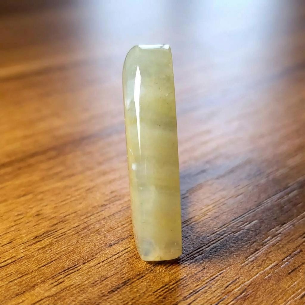 “A vertical standing angle of the polished teardrop-shaped chalcedony agate against a richly grained wooden background. The agate shows layered pale chalcedony with subtle yellow mineral inclusions from old volcanic sediment. The light reflection emphasizes its smooth glossy finish and internal banding.”