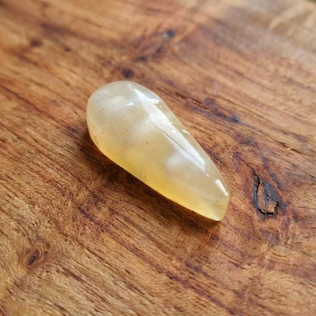 “A side view of the teardrop-cut chalcedony agate standing upright on a wood background. The translucency reveals pale layered chalcedony with muted yellow inclusions and subtle internal banding. Light catches the curved polish, highlighting the trapped volcanic sediment typical of Pacific Northwest volcanic agates.”