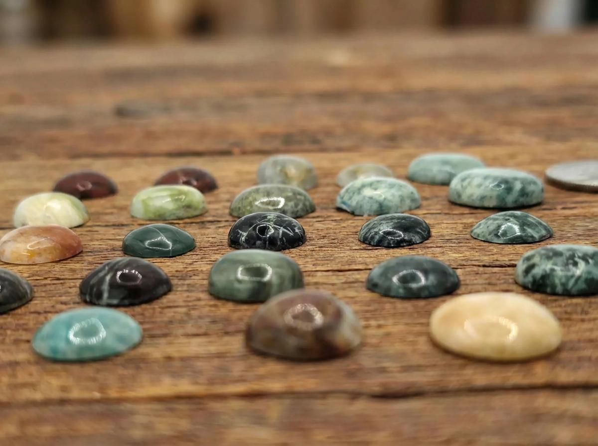 Close-up of polished cabochon gemstones on a wood background, showing detailed textures. Includes green, brown, black, and cream stones with banding, mossy patterns, and orbicular structures. Dome shapes catch overhead light reflections and highlight natural inclusions.